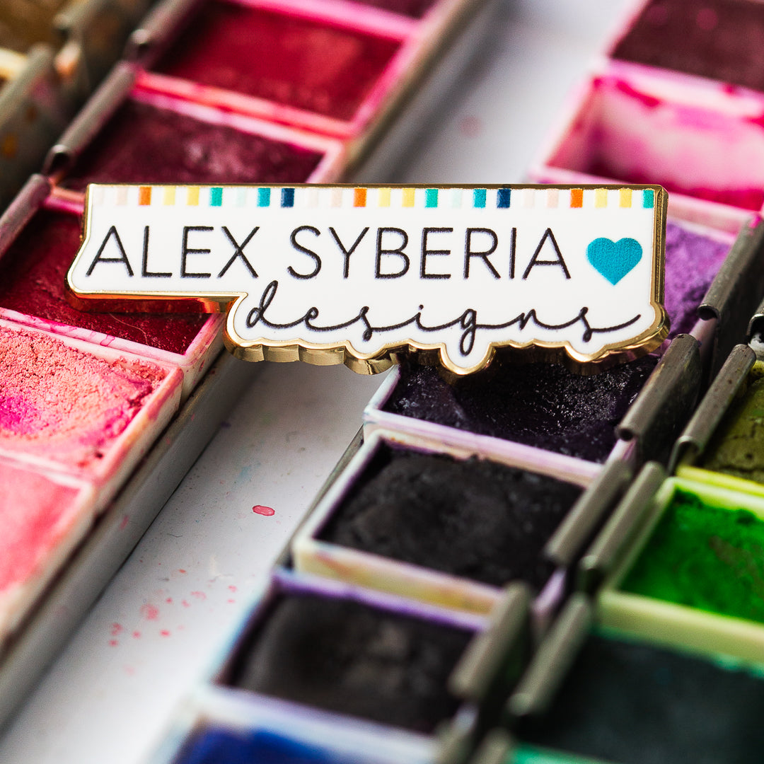 Colorful watercolor paint set with 'ALEX SYBERIA designs' logo enamel pin