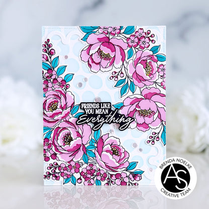 A handmade greeting card featuring a floral design with pink peonies, teal leaves, and a central die-cut sticker that reads 'FRIENDS LIKE YOU MEAN Everything'. The card is displayed on a reflective white surface with a soft, blurred white floral background. Alex Syberia Designs Cardmaking pressplates