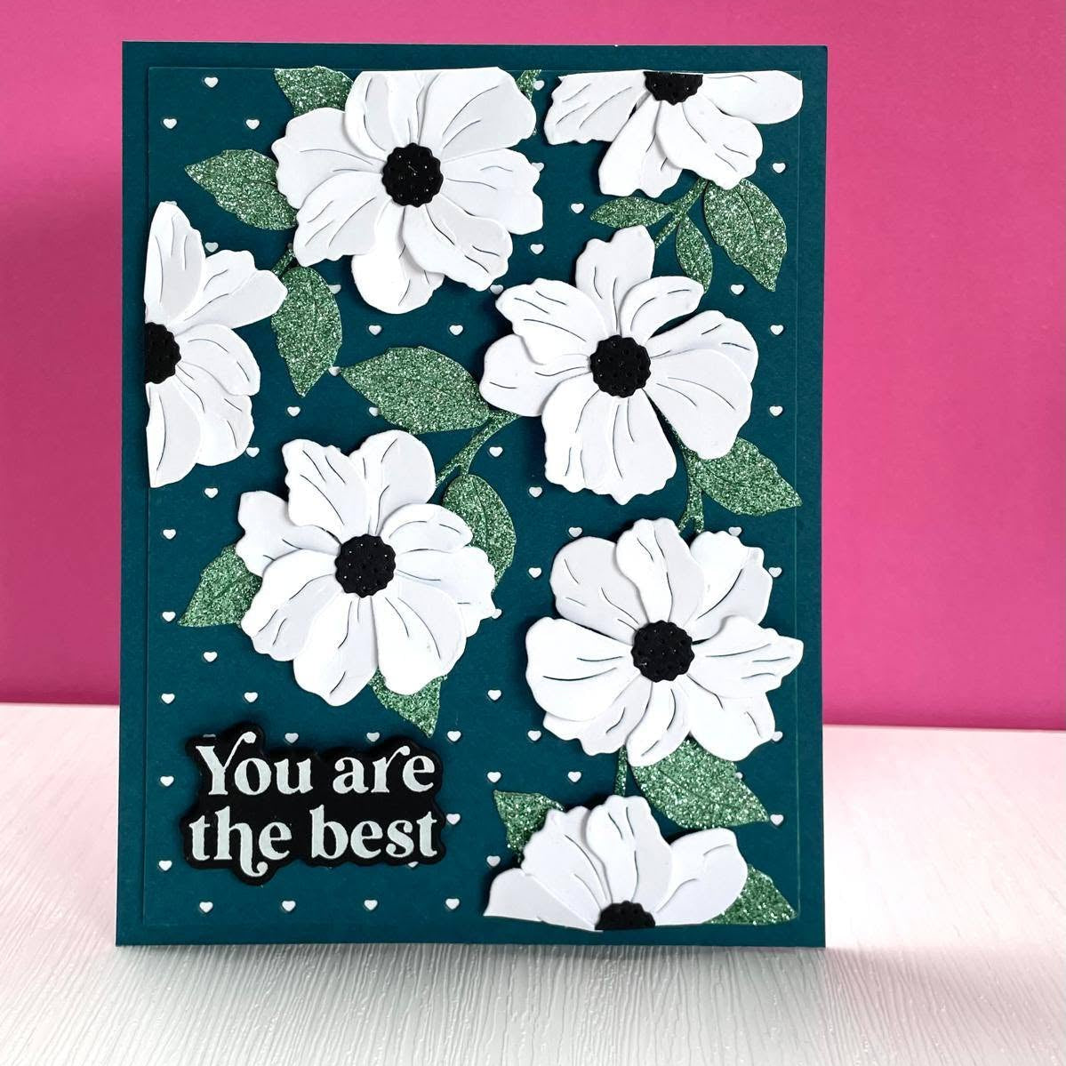 A handmade greeting card with a dark teal background, featuring layered white paper flowers with black centers, green textured leaves, small white hearts, and the text 'You are the best'  Alex Syberia Designs dies in white on a black plaque. The card is displayed on a white surface against a vibrant pink background.