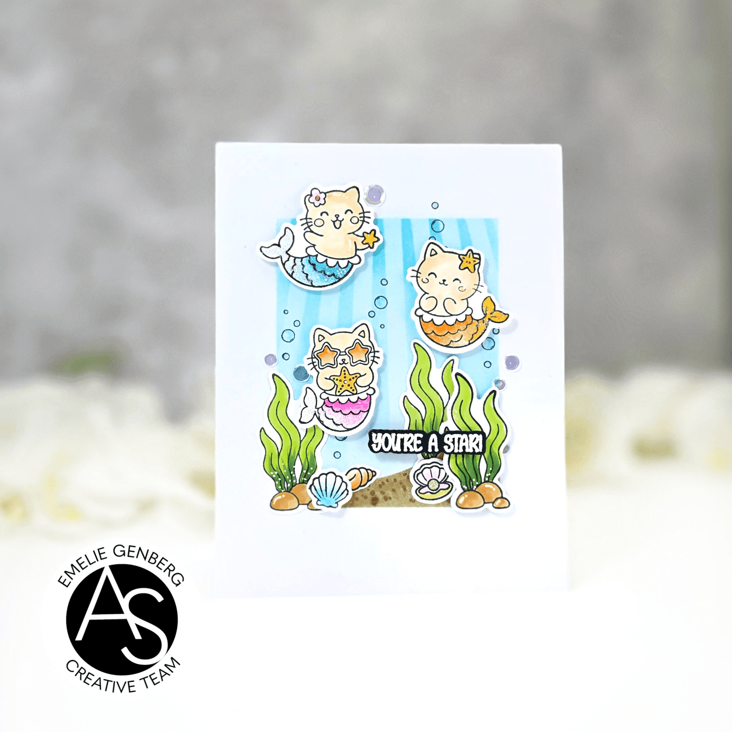 A white greeting card featuring a cute cartoon mermaid cat illustration with the text 'YOU'RE A STAR!' and underwater-themed details, shown against a soft blurred gray and white background. A circular logo in the lower left reads ALEX SYBERIA STAMPS DIES
