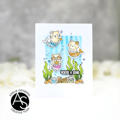 A white greeting card featuring a cute cartoon mermaid cat illustration with the text 'YOU'RE A STAR!' and underwater-themed details, shown against a soft blurred gray and white background. A circular logo in the lower left reads ALEX SYBERIA STAMPS DIES
